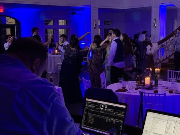 Wedding guests dancing under blue uplighting at The Springs in Weatherford, with the DJ booth and bride visible in the reception ballroom.